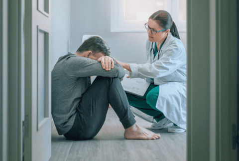 Female doctor reassuring and helping with empathy to male patient sitting on the room floor of a mental health center